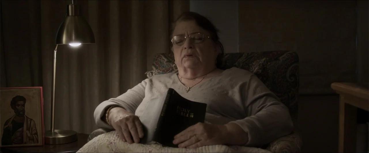 Heavenly Deposit (2019) movie still: medium — A dimly lit room features a patterned armchair and a metal desk lamp. An elderly woman sits in the…