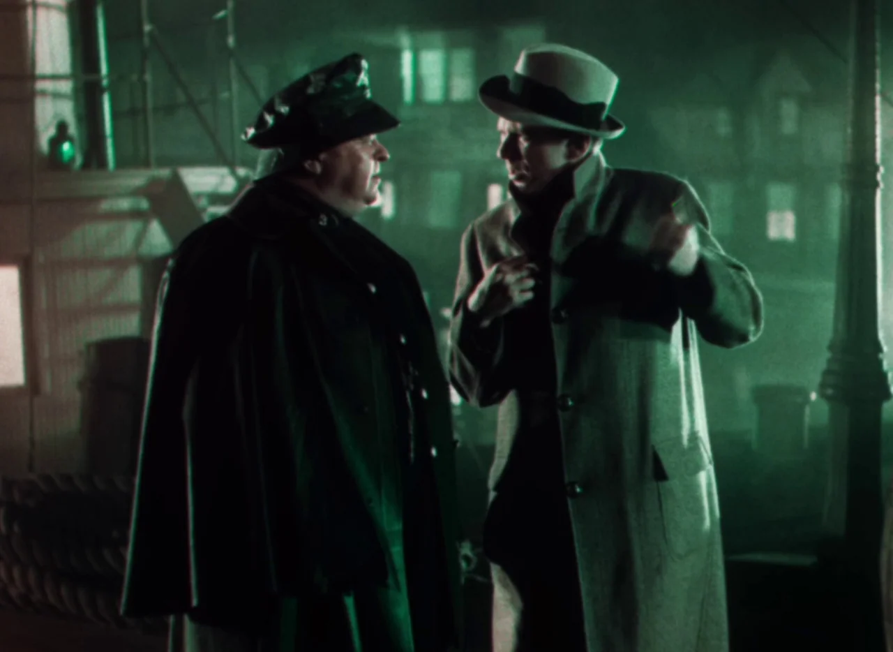 Doctor X (1932) movie still: medium — A night scene on a waterfront with blurred industrial building lights in the distance. Two men in coats and…