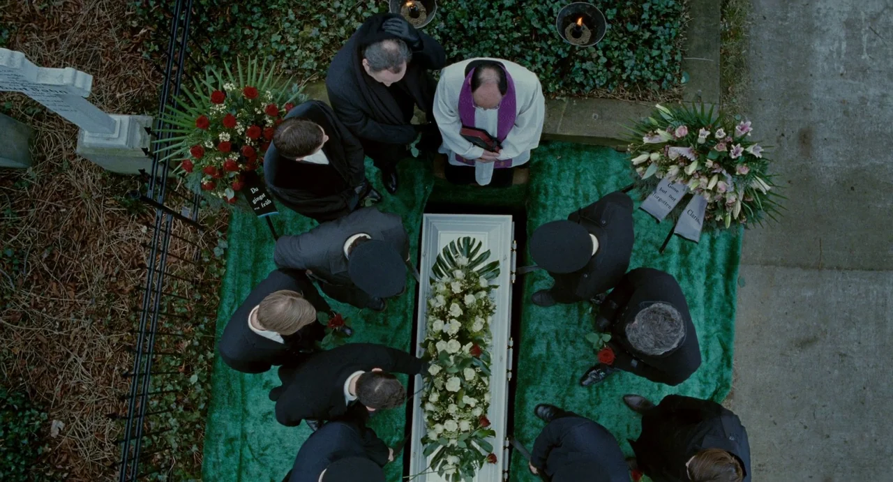 Passion (2012) movie still: wide — An outdoor burial service at a grave covered with synthetic green grass. A group of people dressed in black suits…