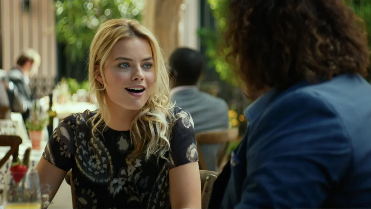 Focus (2015) movie still: medium — An outdoor restaurant seating area features tables with wooden chairs under dappled sunlight. A blonde woman…