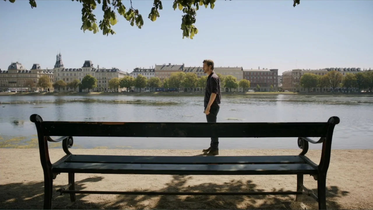 Copenhagen (2002) movie still: fullbody — A park path borders a large calm lake with urban buildings in the distance. A man stands on the dirt path…