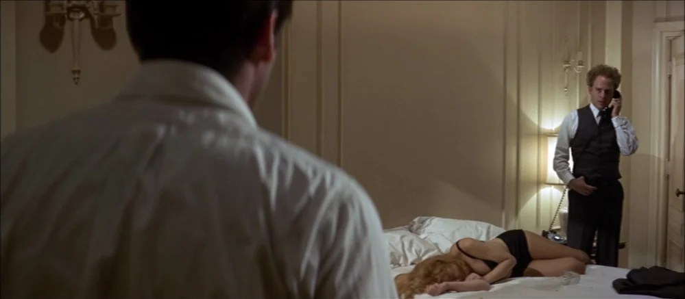 Carnal Knowledge (1971) movie still: medium — A dimly lit hotel room with neutral walls and traditional wall sconces. A man speaks on a telephone…