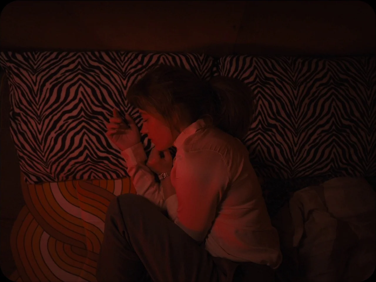 Longlegs (2024) movie still: medium — A person lies on a bed with zebra-print pillows in a dimly lit space. The subject rests in a fetal position…
