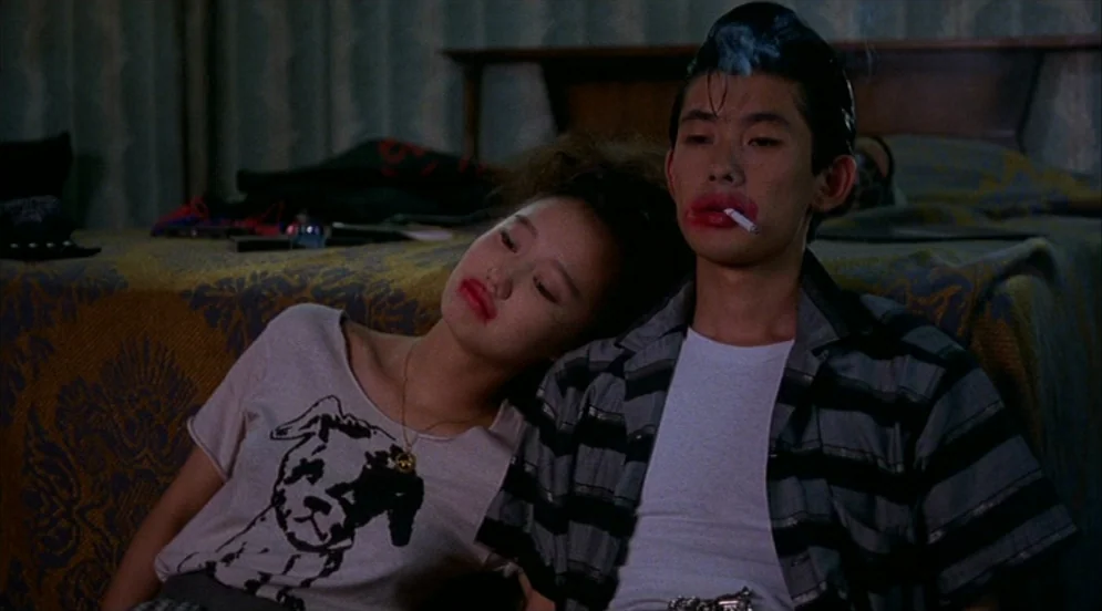 Mystery Train (1989) movie still: medium — A dimly lit room features a rumpled bed with a patterned gold-toned blanket. A young couple sits together…