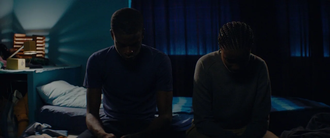 Girlhood (2014) movie still: medium — A dimly lit room with a pair of people sitting on the edge of a bed. The man and woman sit with their backs…