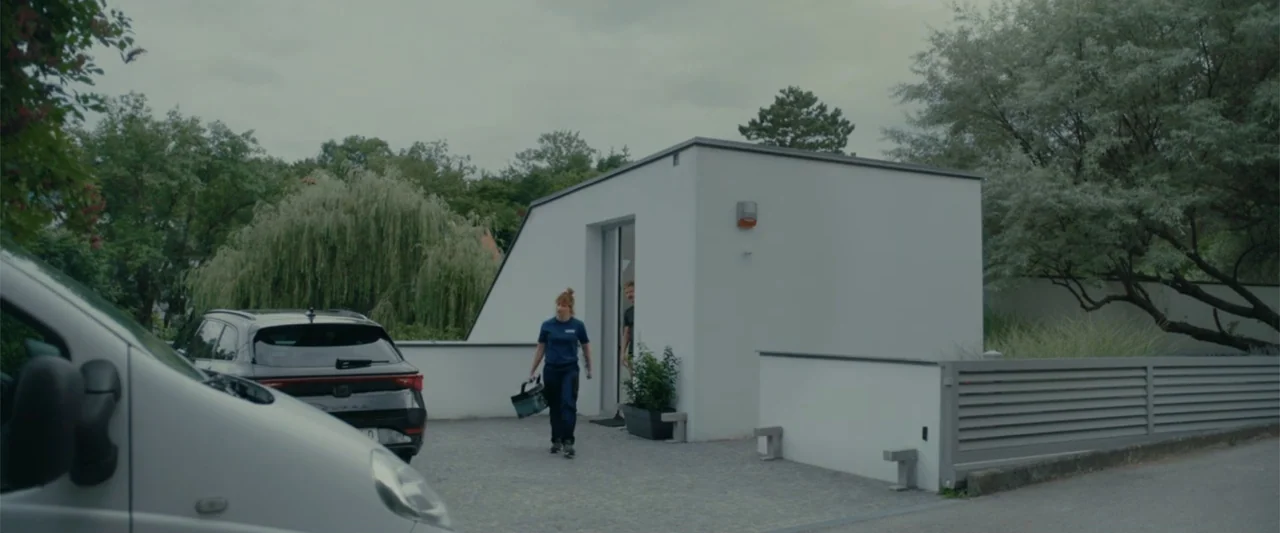 Peacock (2010) movie still: fullbody — A modern white minimalist structure sits behind a grey metal fence on a driveway. A woman in a blue uniform…