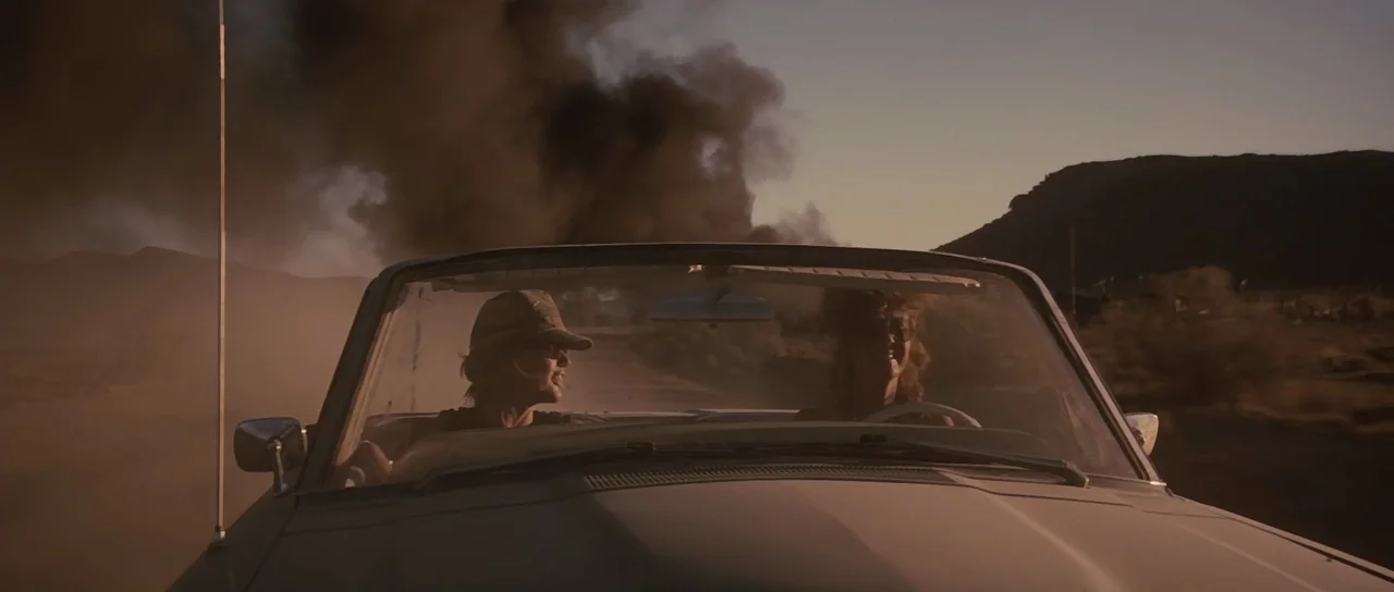 Thelma & Louise (1991) movie still: medium — A convertible car drives through a vast, dry desert landscape with plumes of black smoke. Two women sit…