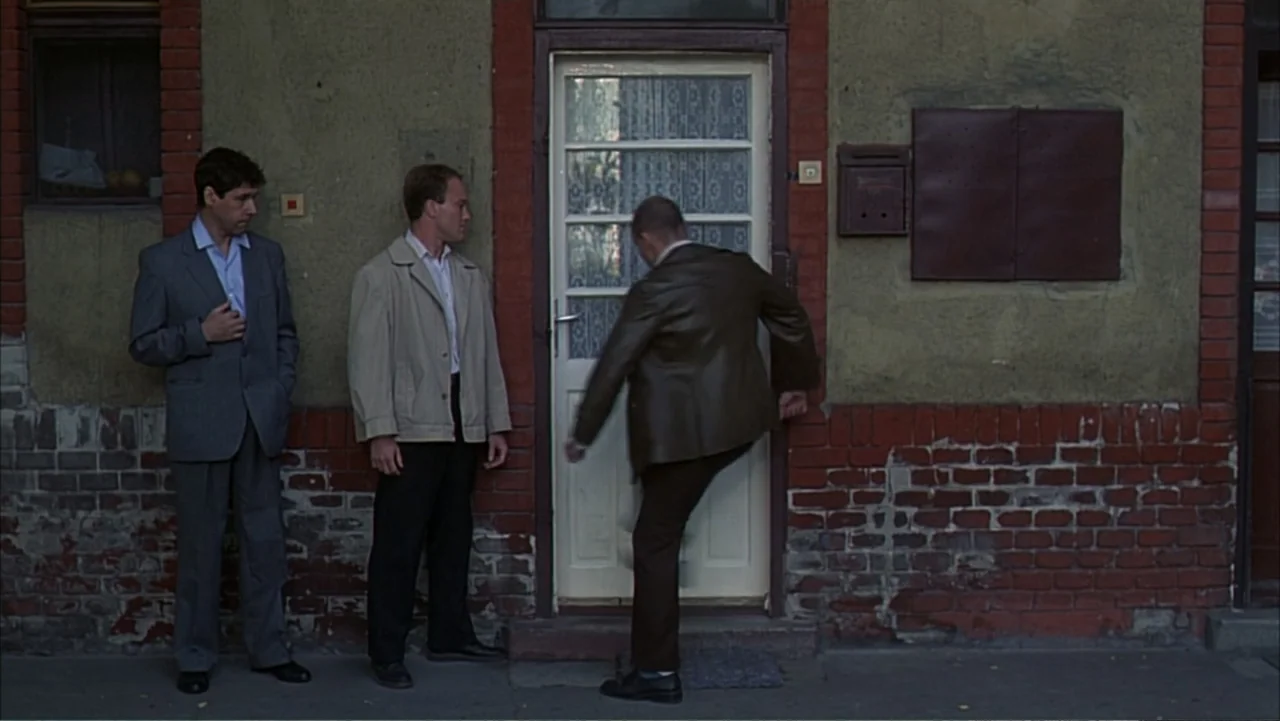 Citizen X (1995) movie still: medium — An exterior wall of an aged building features brickwork and a white entry door. A man kicks the door while…