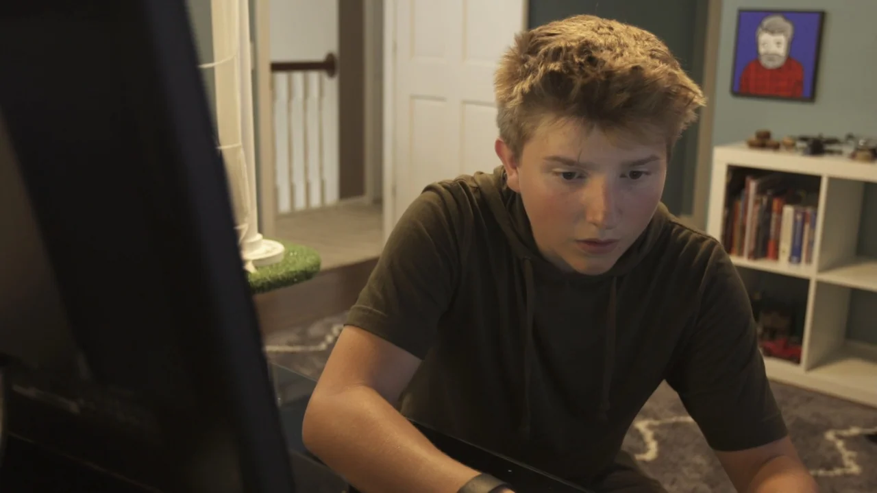 Sk8 Dawg (2018) movie still: medium — A teenager sits in front of a computer monitor in a sparsely decorated room. The boy looks down at his…