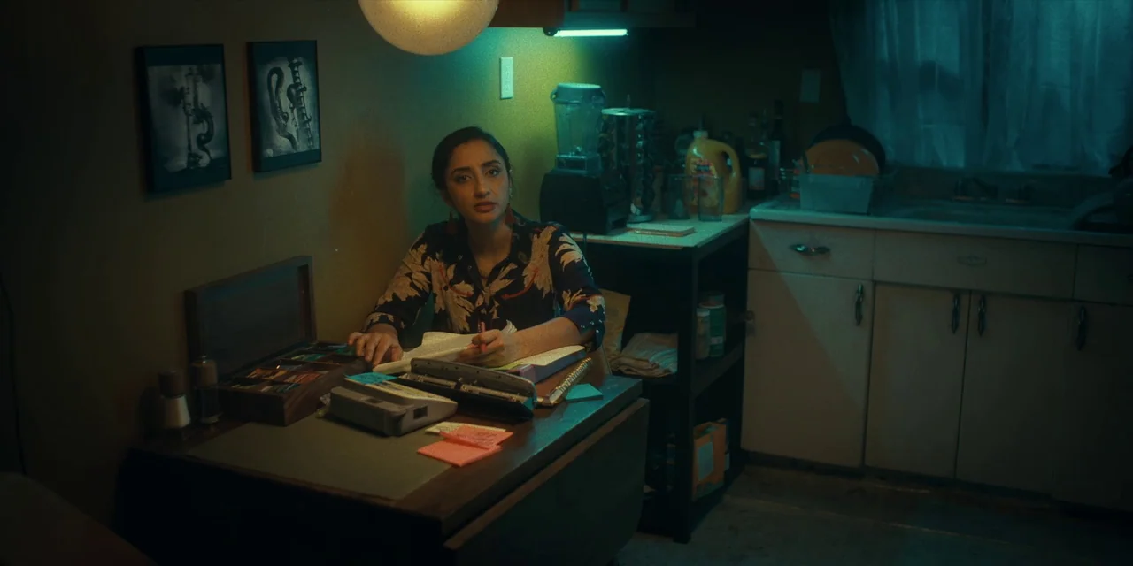 Destroy All Neighbours (2024) movie still: medium — A woman sits at a wooden desk cluttered with papers and a staple gun. The room behind her has…