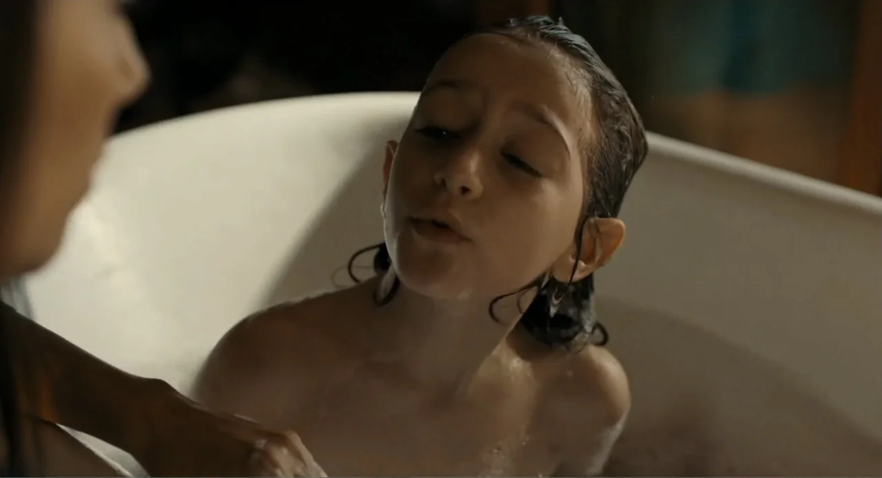 Elles (2018) movie still: closeup — A white bathtub contains soapy water and a young child with wet hair. An adult stands out of focus in the…