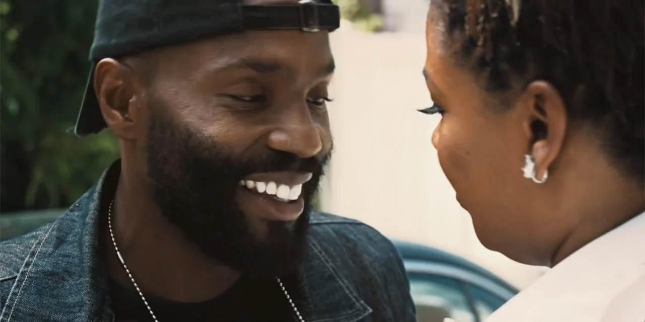 Equal Standard (2021) movie still: closeup — An outdoor parking area features a light-colored vehicle blurred in the background. A man smiles while…