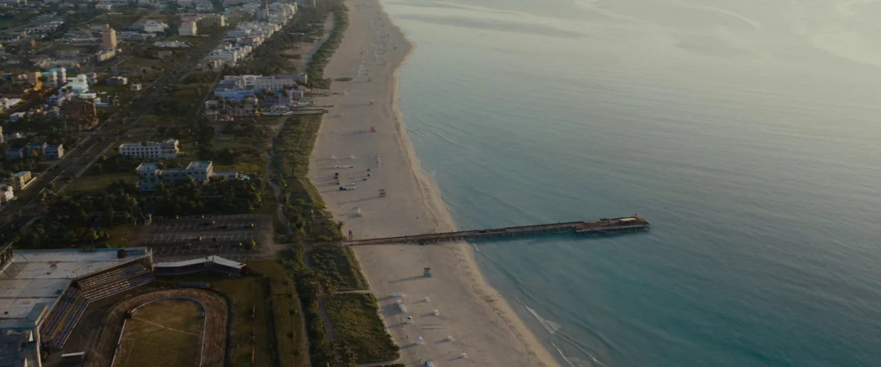 Live By Night (2016) movie still: establishing — An aerial view shows a long stretch of sand meeting the ocean with a pier. A coastal city with…