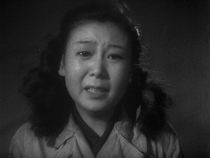 One Wonderful Sunday (1947) movie still: closeup — A dark space serves as the background for a person in a trench coat. The woman faces the camera…