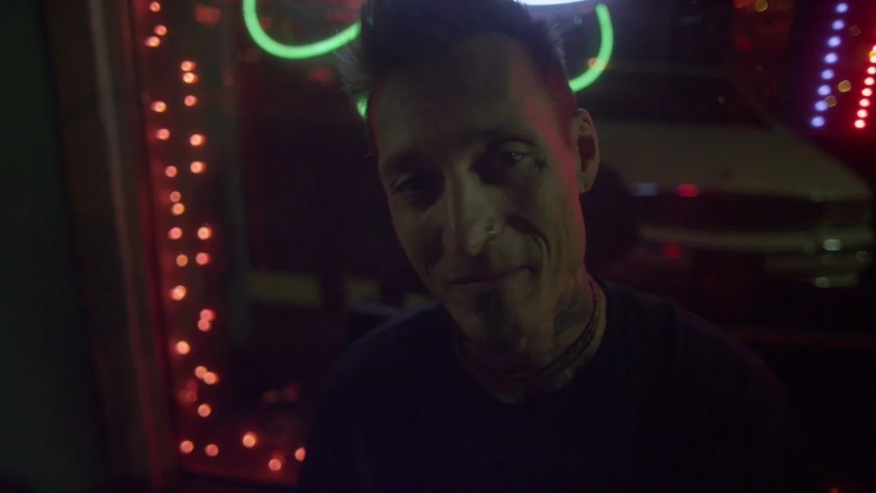 Florida Man (2015) movie still: closeup — A man stands outdoors at night near bright neon signage reflecting on his face. The man maintains a steady…