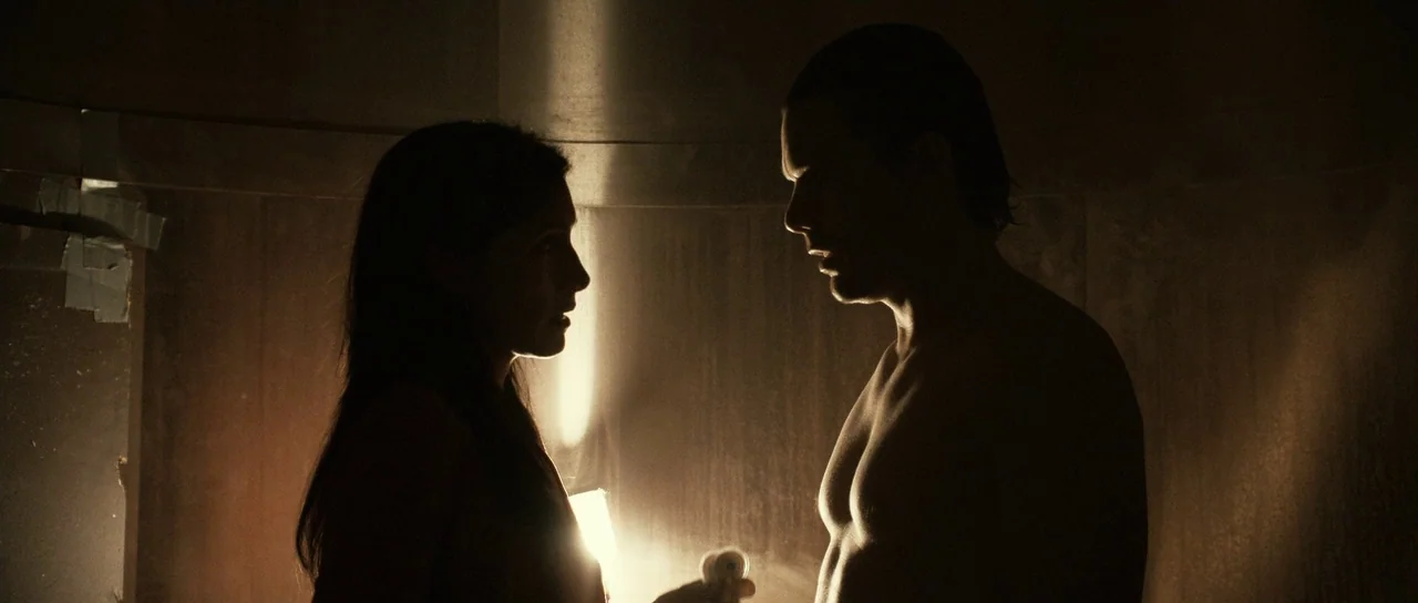 Daybreakers (2009) movie still: medium — A dimly lit interior space features two figures silhouetted against a singular bright light. The man and…
