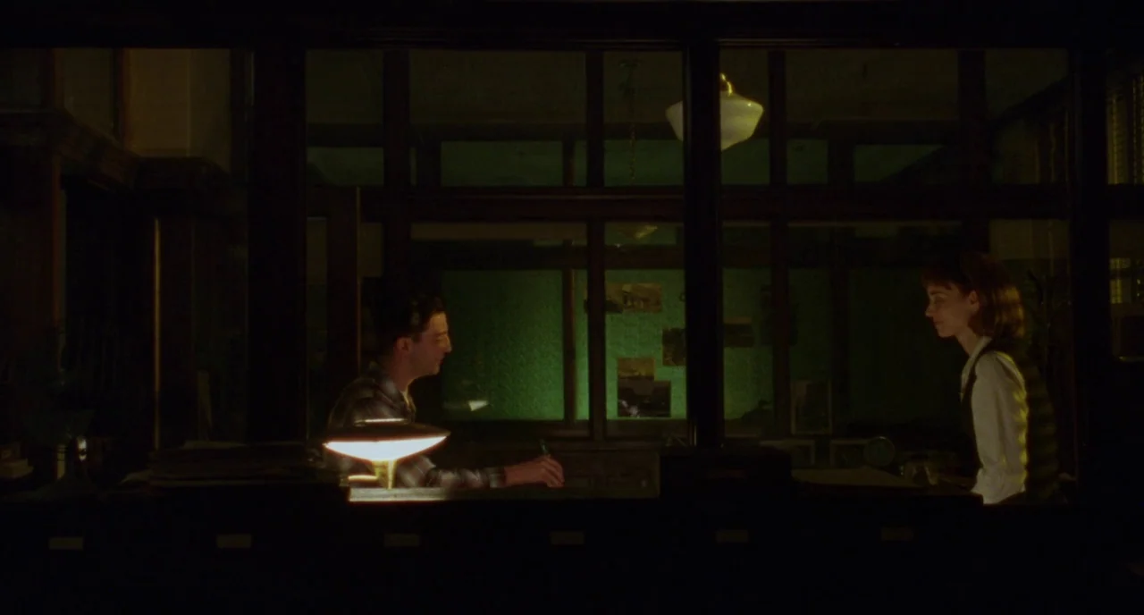 Carol (2015) movie still: wide — A dimly lit room features a large window divided by dark wooden muntins. A man and woman occupy the workspace,…