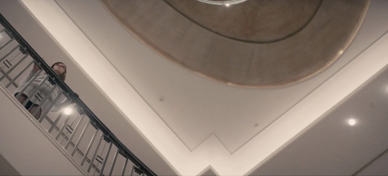 Housewife (2017) movie still: wide — A bright multi-level interior features a white staircase railing and a large curved ceiling. A woman stands on…