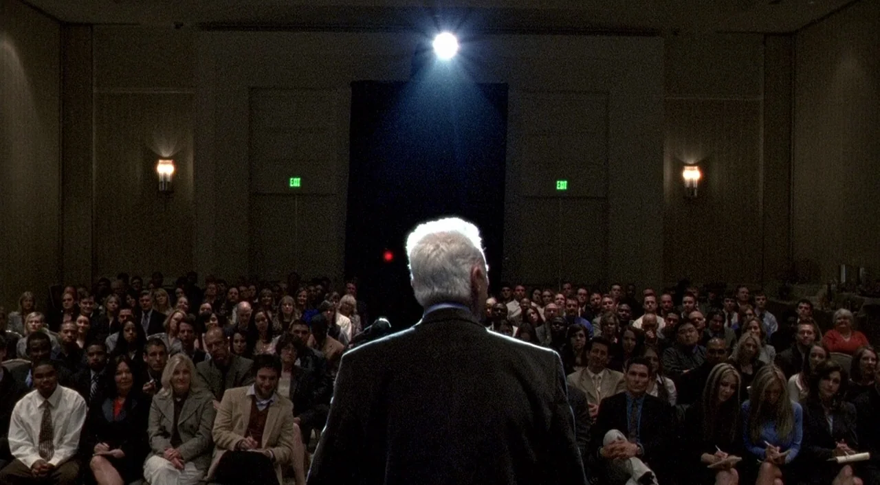 Halloween 2 (2009) movie still: medium — A dark, dimly lit conference hall is filled with a large crowd of people seated in rows. A man in a dark…