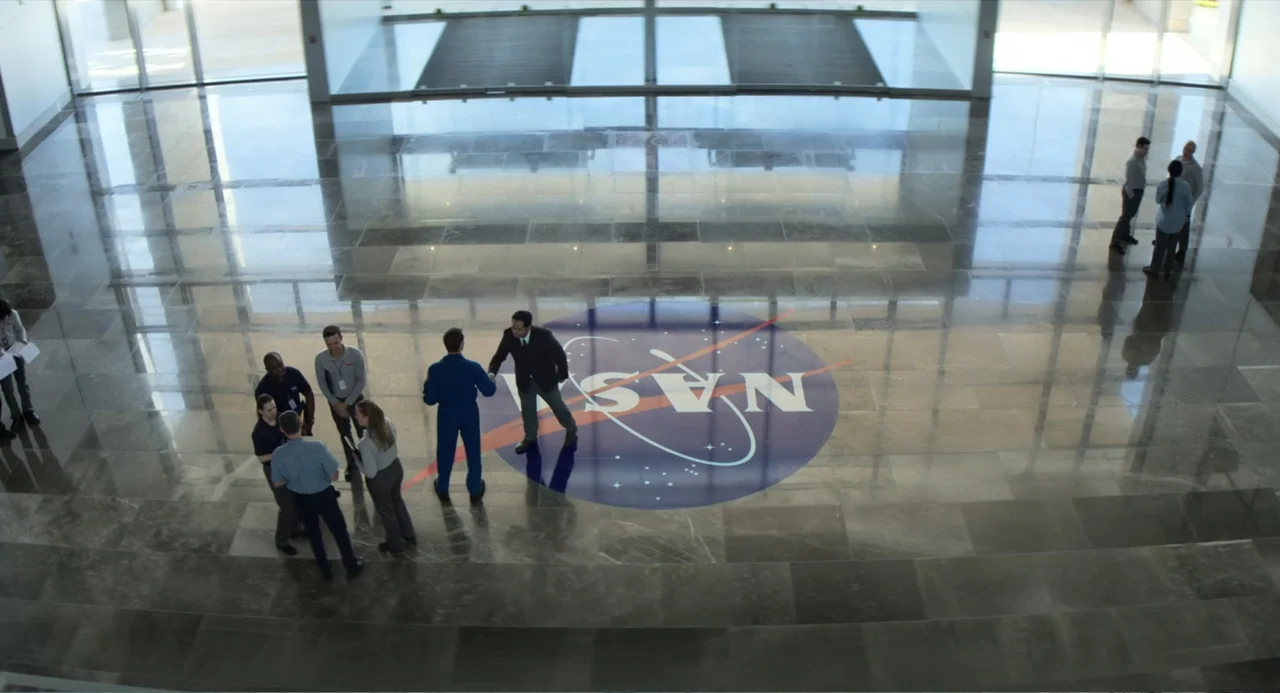 A Million Miles Away (2023) movie still: wide — A spacious government building lobby features a large NASA logo inlaid on the floor. A group of…