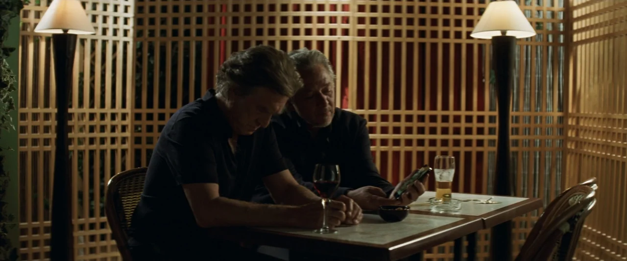 Masquerade (2022) movie still: medium — A restaurant booth features a wooden lattice screen wall behind two seated men. The men sit closely together…