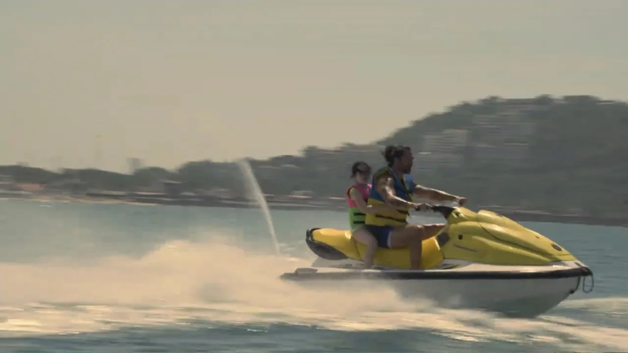 Double Pregnant 2 (2022) movie still: medium — A man and a child ride a yellow jet ski across open coastal water. The shoreline with buildings and…