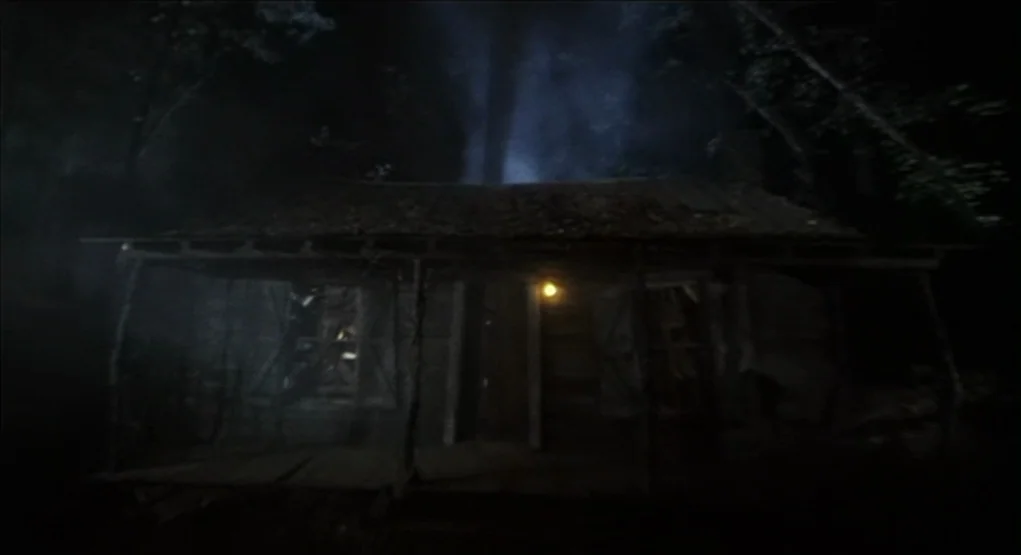 Evil Dead II (1987) movie still: establishing — A weathered wooden cabin stands at night surrounded by tall trees and thick fog. The structure…
