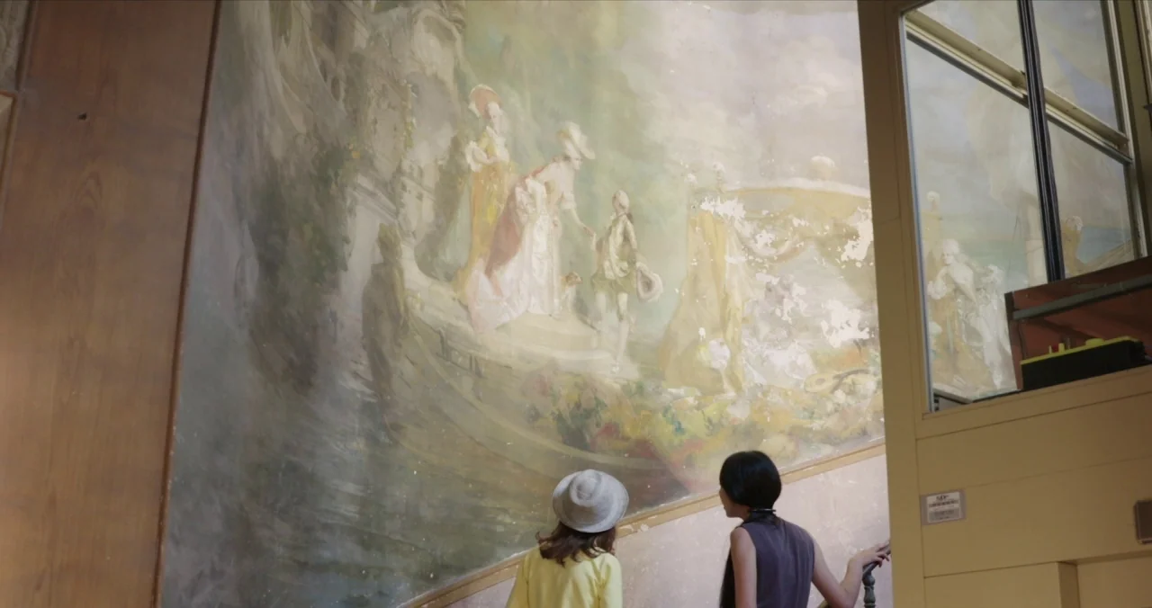 Claire’s Camera (2017) movie still: medium — Two women stand in a stairwell looking at an expansive 18th-century style wall mural. The painted scene…