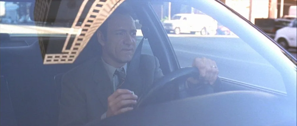 American Beauty (1999) movie still: medium — A man drives a vehicle on a bright, sunny city street during the day. He holds a cigarette while…