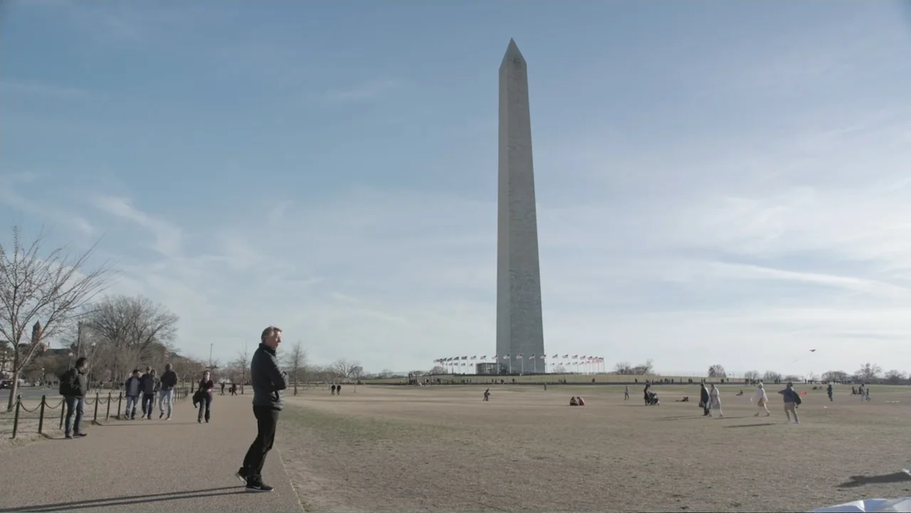 A Gift from God (2019) movie still: wide — The Washington Monument stands prominently in a large open park under a clear sky. A man in a dark jacket…