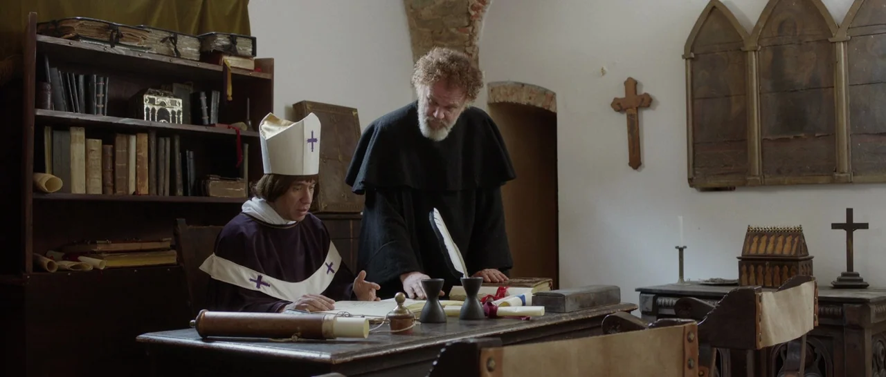 The Little Hours (2017) movie still: medium — A stone-walled study features wooden bookshelves filled with aged scrolls and thick volumes. A bishop…
