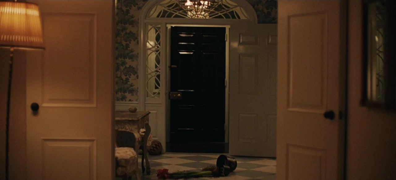 Babygirl (2024) movie still: wide — A dimly lit interior foyer features patterned wallpaper and a black front door. An overturned terracotta pot…