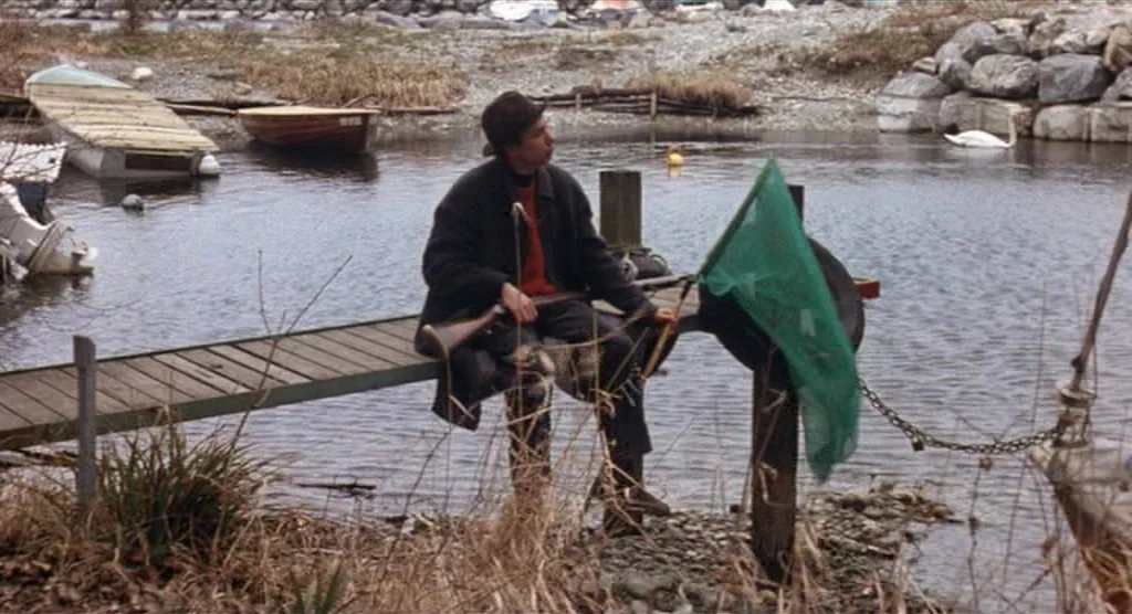 King Lear (1987) movie still: fullbody — A man sits on a narrow wooden dock extending into a gray river. Several small boats and a swan are visible…
