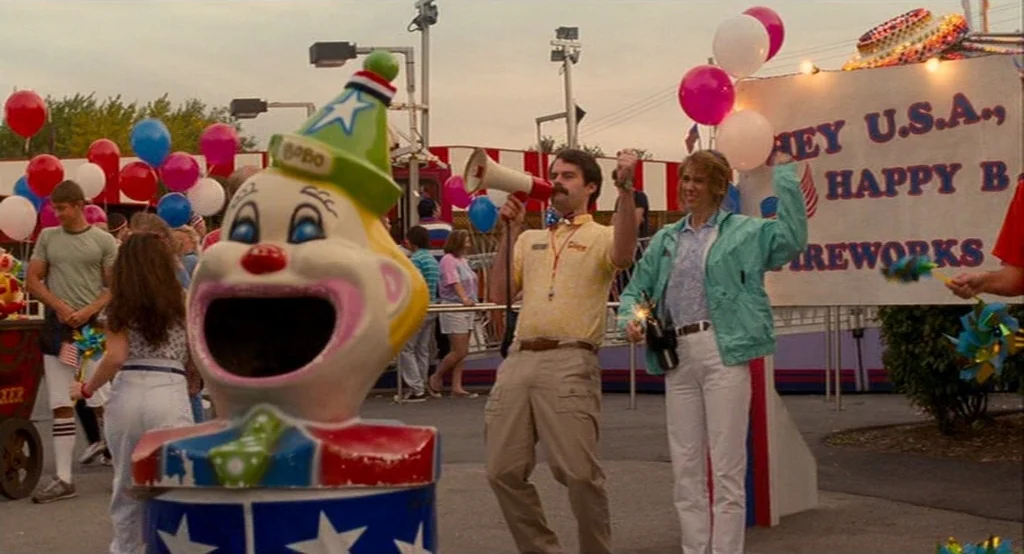 Adventureland (2009) movie still: medium — An outdoor carnival setting featuring a large clown-themed trash receptacle in the foreground. A man with…
