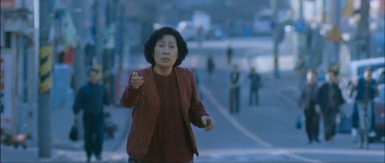Mother (2009) movie still: medium — A quiet urban street features blurred buildings and pedestrians moving in the distance. The woman remains…