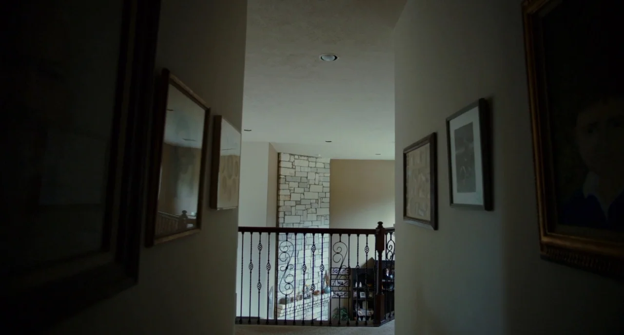 Krisha (2015) movie still: establishing — A narrow residential hallway features beige walls adorned with framed rectangular pictures on both sides.…