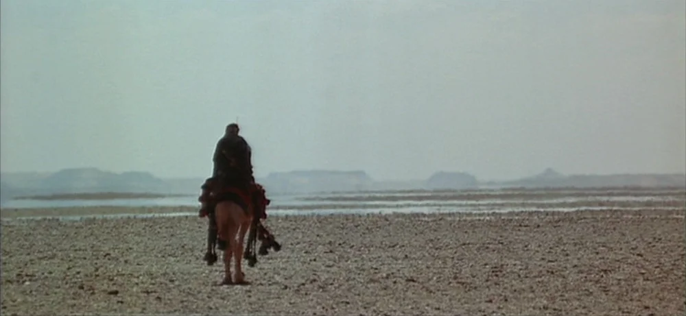 Lawrence Of Arabia movie still: wide — A vast, flat, rocky desert floor stretches toward distant, low-lying hills under pale skies. A lone figure…
