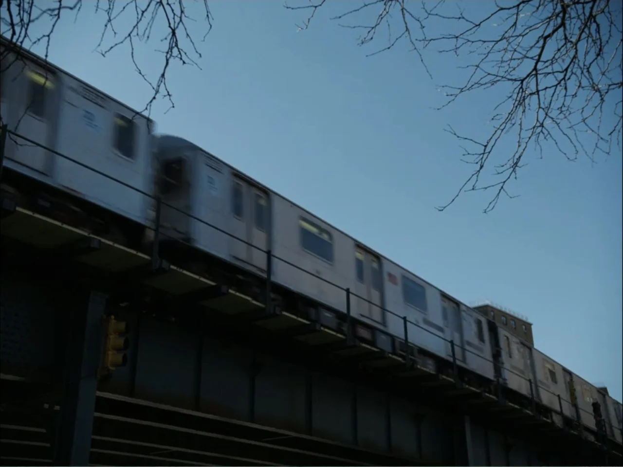 As We Speak: Rap Music on Trial (2024) movie still: wide — An elevated train traverses a steel bridge against a clear blue sky. Leafless branches…