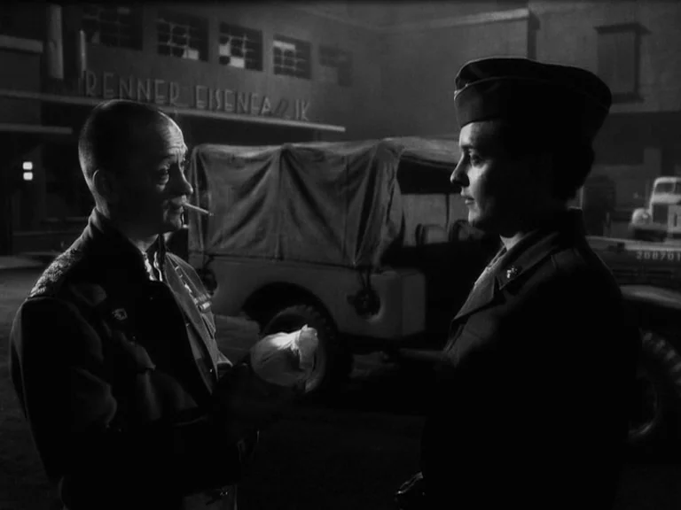 The Good German movie still: medium — A dark industrial site at night features a large warehouse and a parked truck. A man in a uniform talks to a…