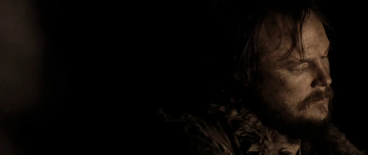 Van Diemens Land (2009) movie still: closeup — A man stands in a dark environment with a fur-lined garment visible. He is looking down while…