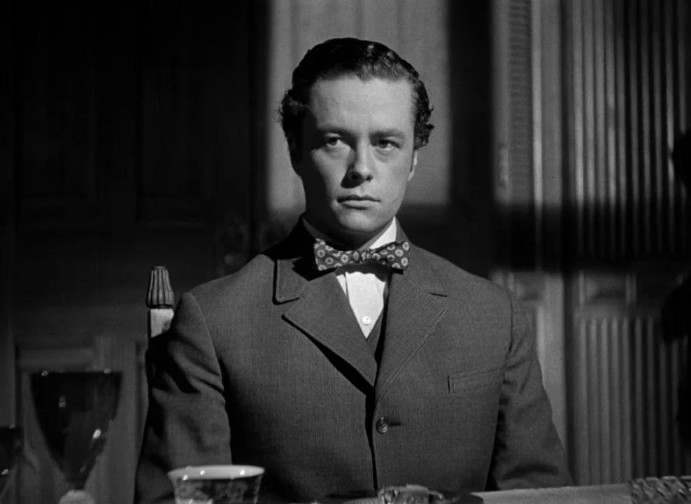 The Magnificent Ambersons (1942) movie still: medium — A man sits at a dining table inside a dark room with paneled walls. He stares forward while…