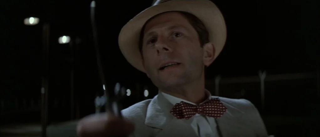 Chinatown (1974) movie still: closeup — A dark outdoor setting at night features blurred lights in the distant background. A man in a suit and hat…