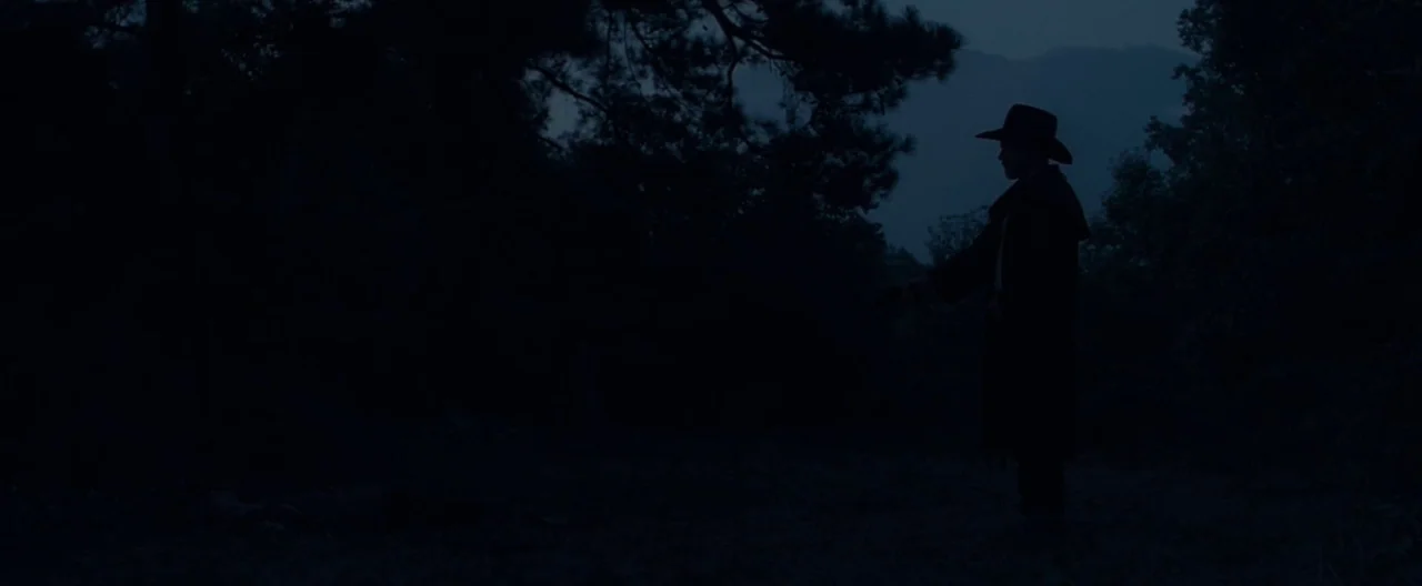 Ain’t Them Bodies Saints (2013) movie still: wide — A dark, wooded area at night is filled with shadows and tall pine trees. A man wearing a duster…