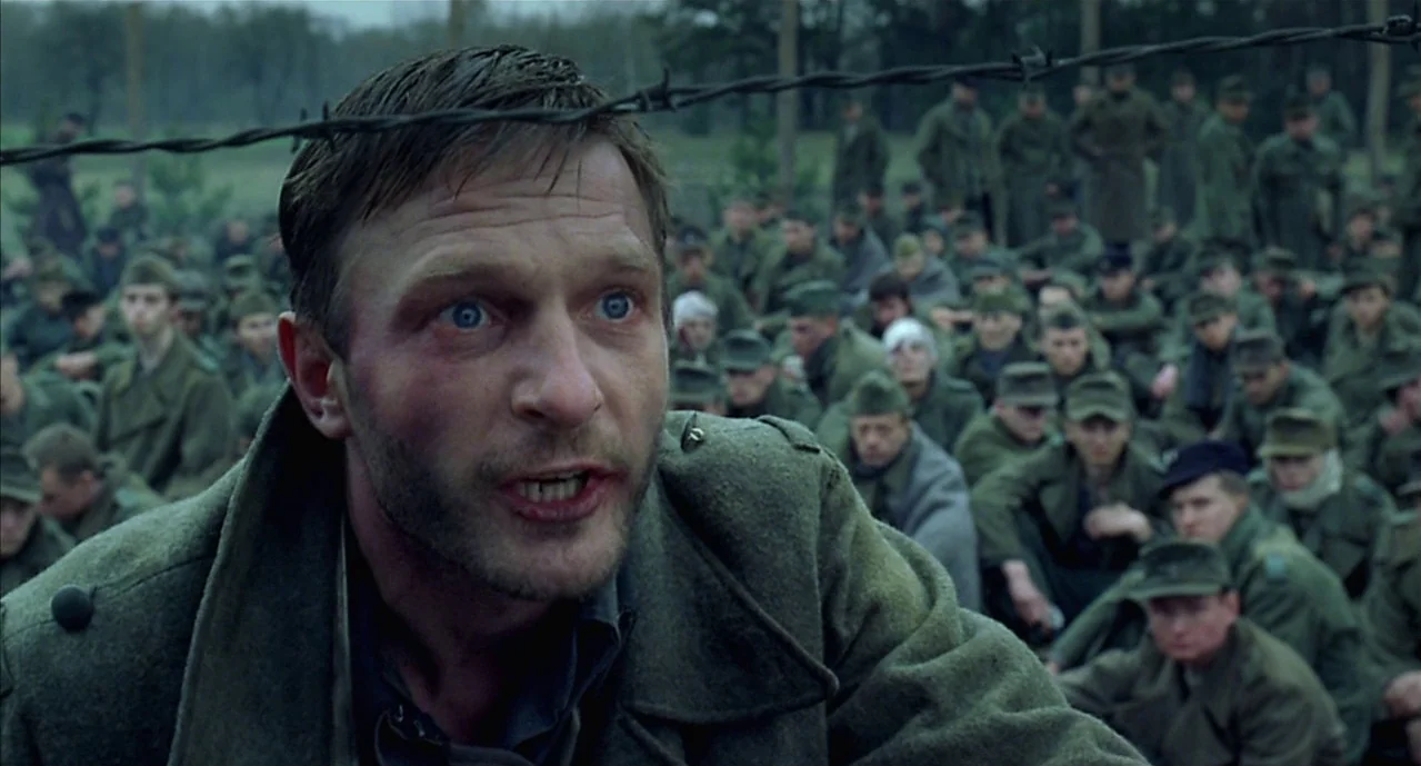 The Pianist (2002) movie still: closeup — An outdoor prisoner of war camp has barbed wire visible in the extreme foreground. A man stands in front…