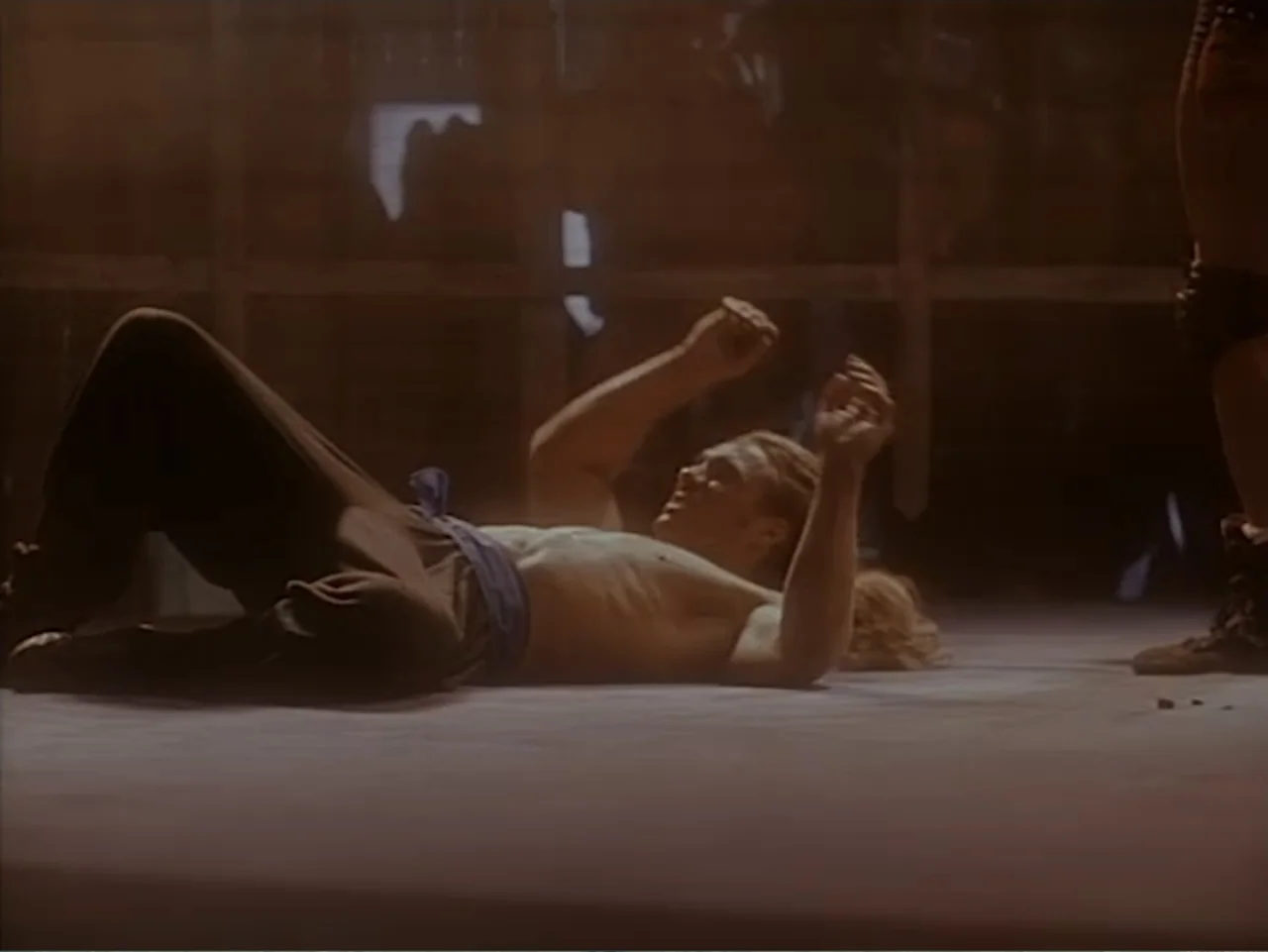 Firepower (1979) movie still: medium — A wrestling ring with wooden ropes and a canvas floor in a dimly lit hall. A man with a bare torso lies on…