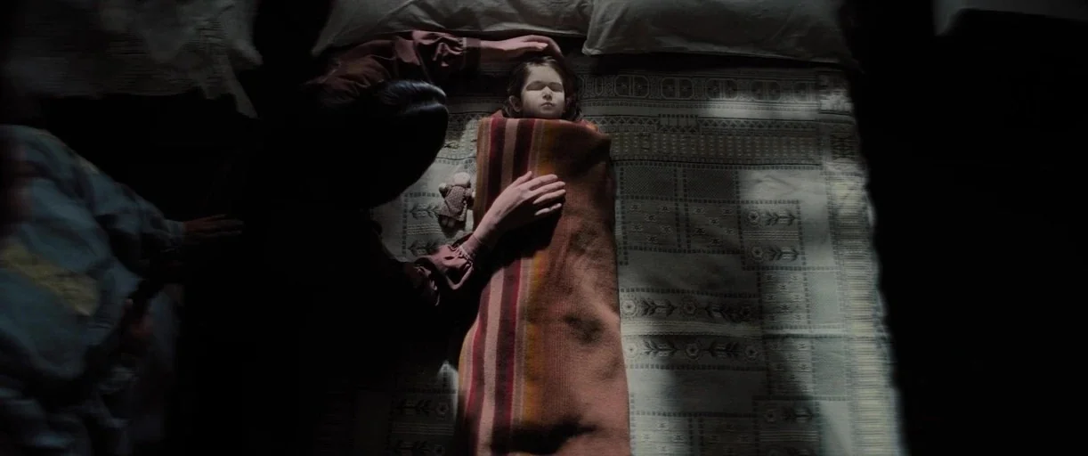 Killers of the Flower Moon (2023) movie still: medium — A top-down view shows a small child lying still under a striped wool blanket. An adult woman…