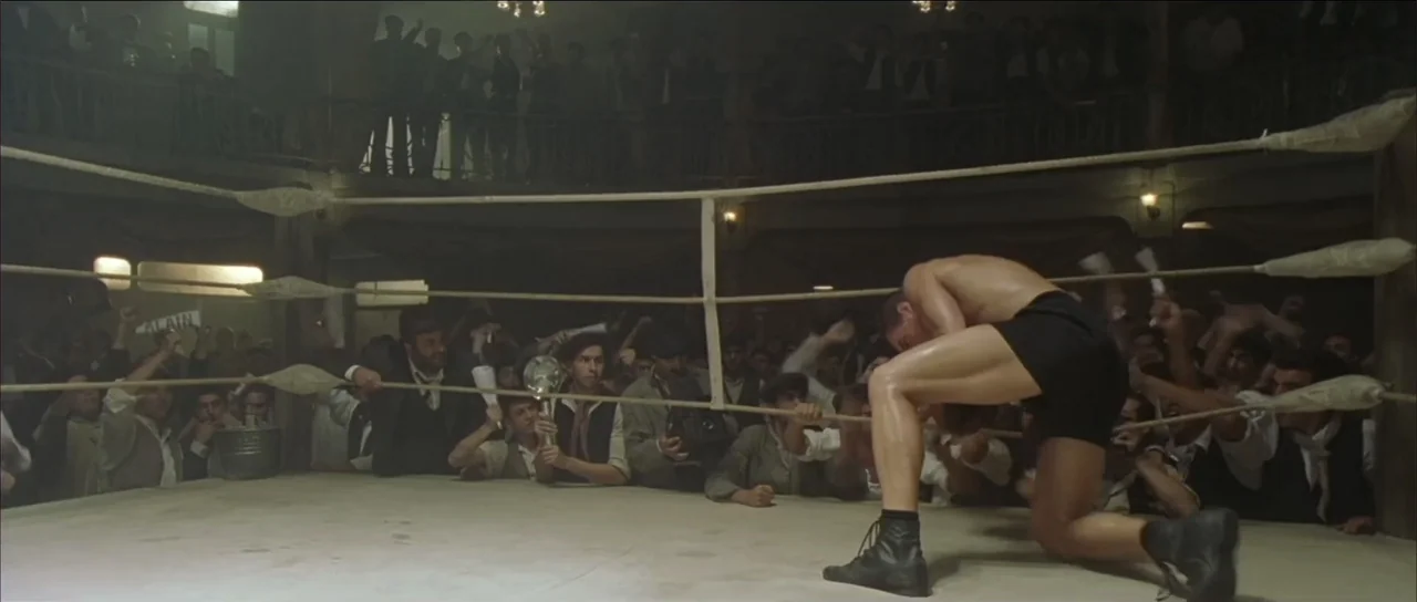 Legionario (2025) movie still: medium — An indoor vintage boxing ring is packed with a large, cheering 1920s crowd. A shirtless muscular boxer in…