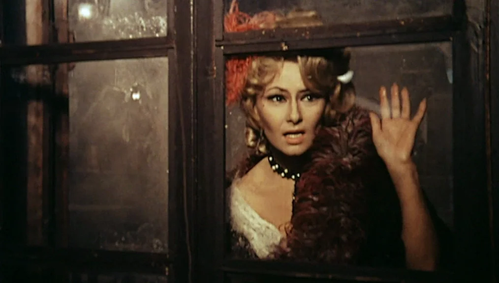 The Great Silence (1968) movie still: medium — A woman stands behind a dark-paned wooden window frame in a dim room. She looks forward with her left…