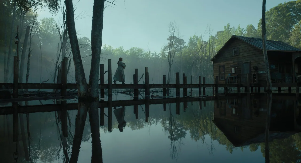 The Color Purple (2023) movie still: wide — A misty, expansive swamp features a weathered wooden pier leading to a small building. A woman in a long…