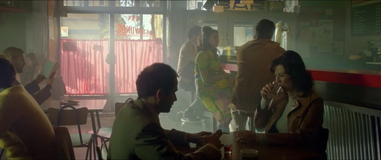 My Heart Goes Boom! (2020) movie still: medium — A smoky 1970s bar filled with patrons sitting at tables and the counter. A man and a woman in the…